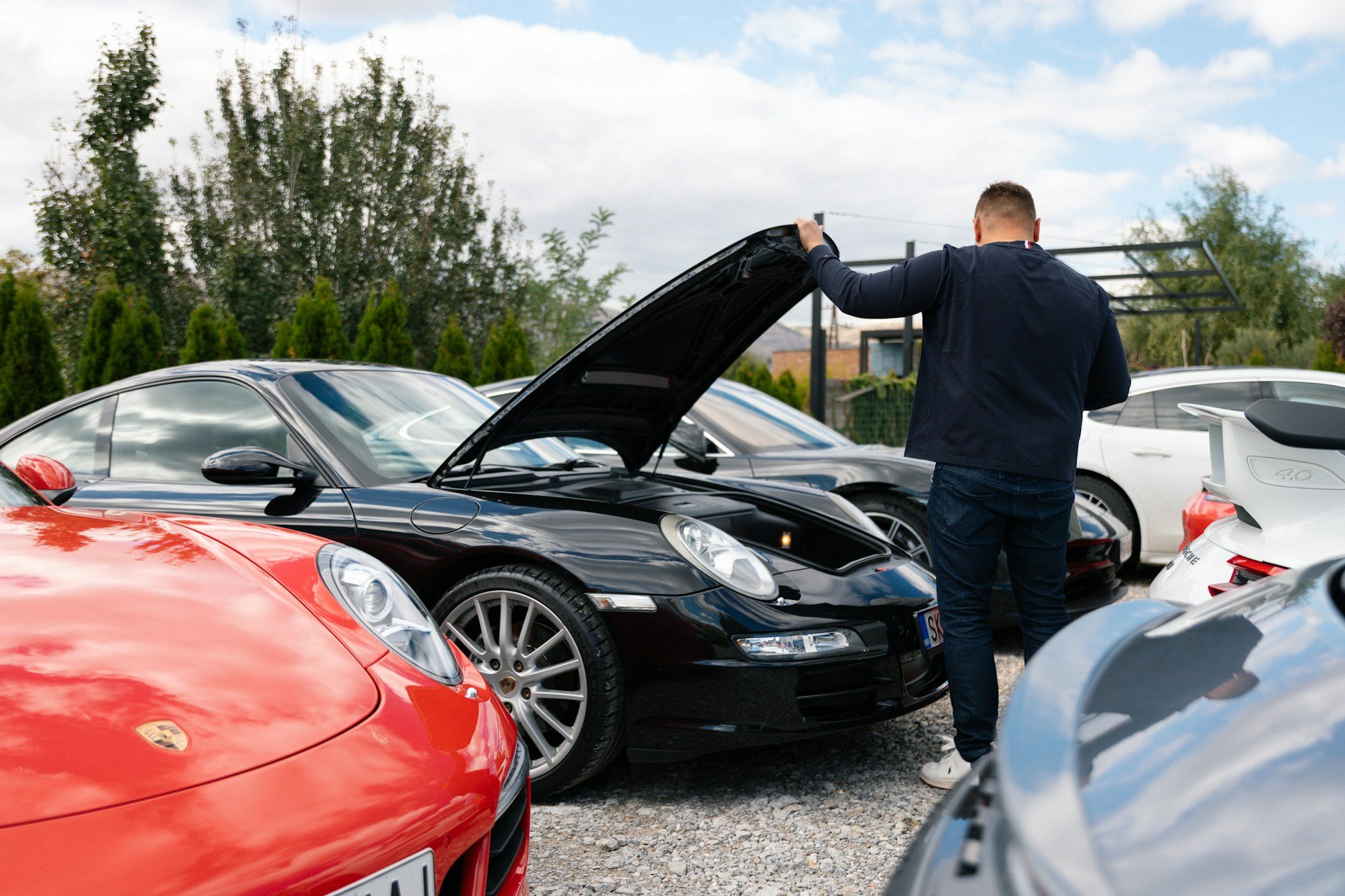 Man inspecting engine of black Porsche 911 surrounded by other sports cars
