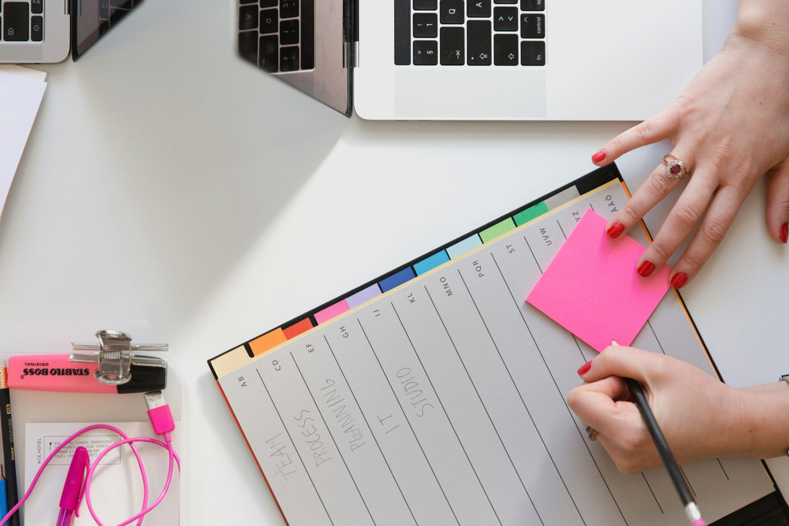 Overhead view of hands writing notes on a pink sticky note and planner near a laptop