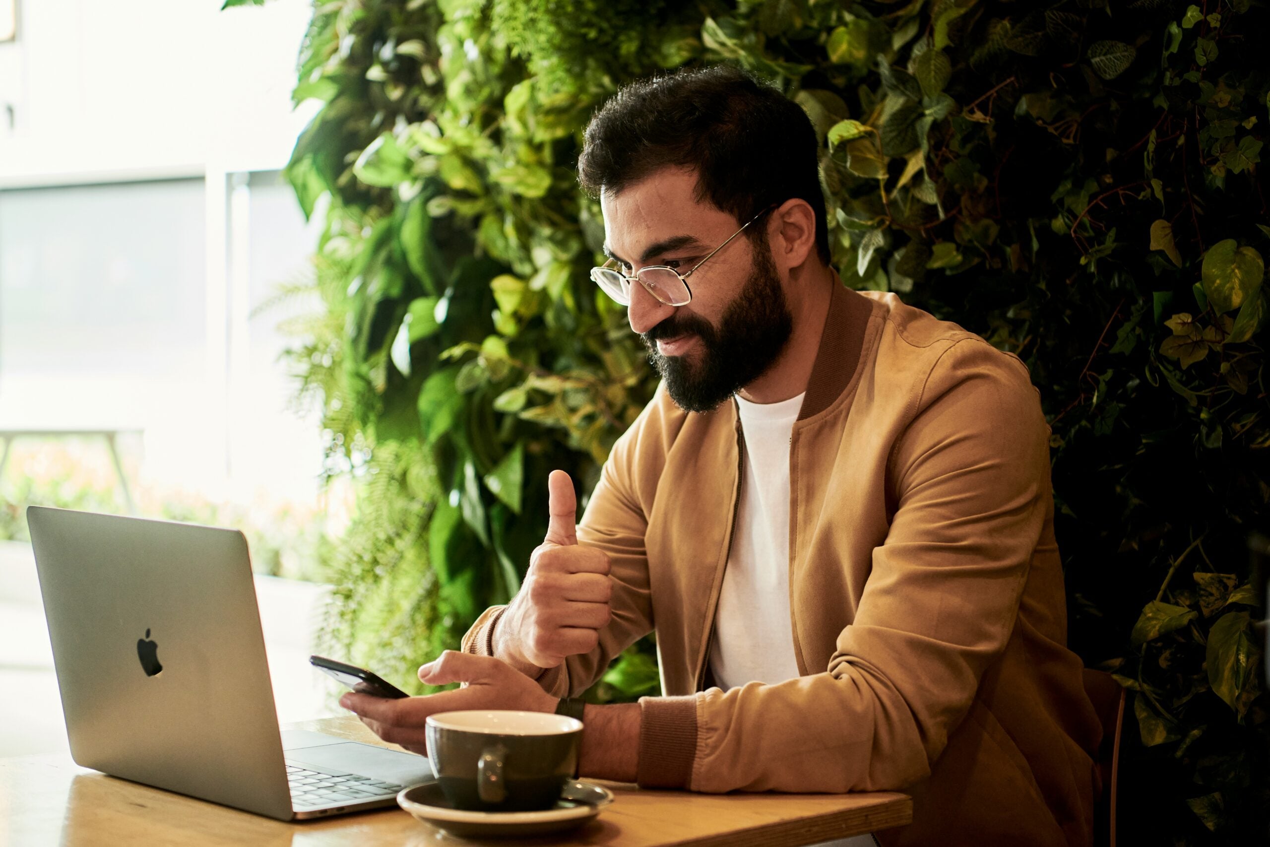 Man with glasses giving a thumbs up while using a laptop and phone